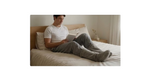 A man relaxes on a bed reading a book, wearing a white t-shirt and gray foot warmer
