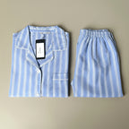 A two-piece pajama set laid flat on a neutral beige background, featuring a light blue and white striped long-sleeve button-up shirt with a collar and matching striped shorts.