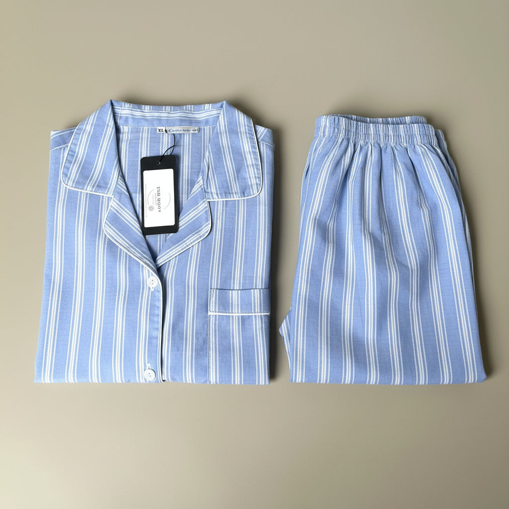 A two-piece pajama set laid flat on a neutral beige background, featuring a light blue and white striped long-sleeve button-up shirt with a collar and matching striped shorts.