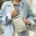 A person wearing a light blue button-up shirt and gray top carries a beige crossbody bag with black trim and a wide shoulder strap.