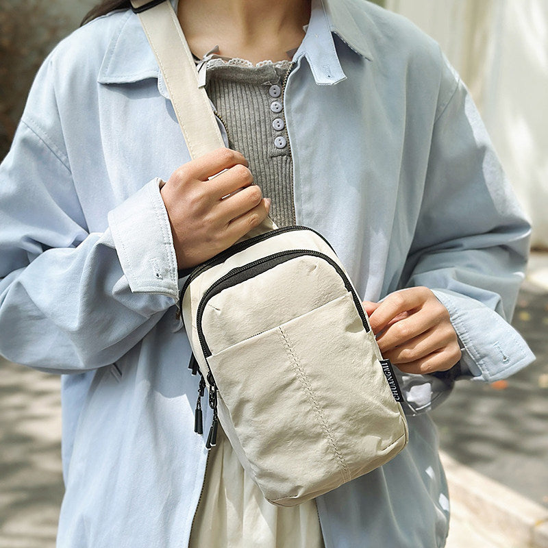 A person wearing a light blue button-up shirt and gray top carries a beige crossbody bag with black trim and a wide shoulder strap.