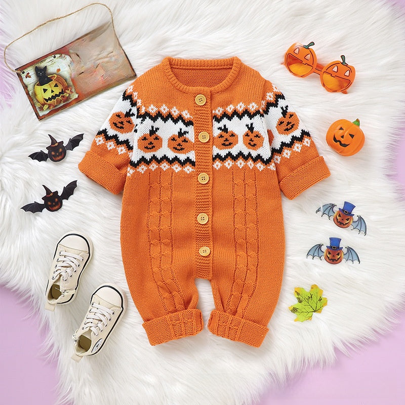 A baby romper laid flat on a white faux-fur surface with Halloween-themed props. The romper is orange with a white yoke pattern featuring black zigzags and orange jack-o'-lantern faces, plus gold-tone buttons.