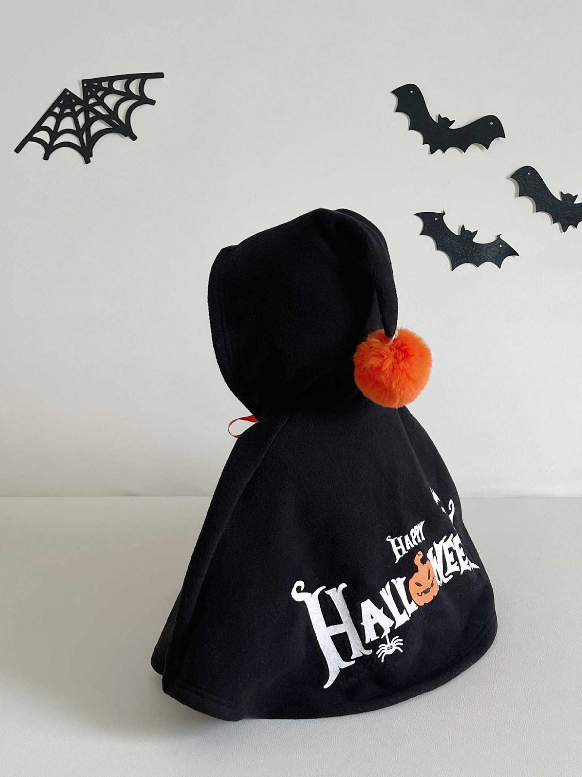 A black hooded cape costume with 'Halloween' printed in white gothic lettering and an orange pumpkin graphic, featuring a small orange pom-pom on the hood, set against a white background with black bat and spiderweb decorations.
