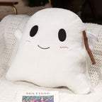 A white plush ghost-shaped pillow with embroidered black eyes and mouth, pink cheek details, and a brown scythe accessory, resting on a white knitted blanket.