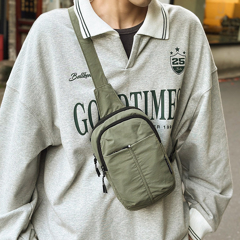 A person wearing an oversized light gray sweatshirt with a green crossbody sling bag worn across the chest. Keep your phone, wallet, keys and other small essentials organised in the zipped compartments.