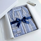 A two-piece pajama set laid flat on a light background, featuring a long-sleeve button-up shirt and matching pants. The fabric has a vertical stripe pattern in shades of blue, navy, brown, and white.