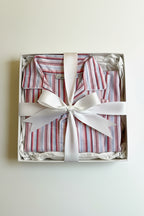 A folded striped pajama shirt presented inside a white gift box with crumpled tissue paper and tied with a wide white ribbon. The pajama has red, pink, and white vertical stripes.