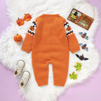 An orange knit baby jumpsuit with Halloween-themed patterns on the sleeves, laid flat on a white faux fur rug with small props including orange glasses, a pumpkin, shoes, and a hanging sign.