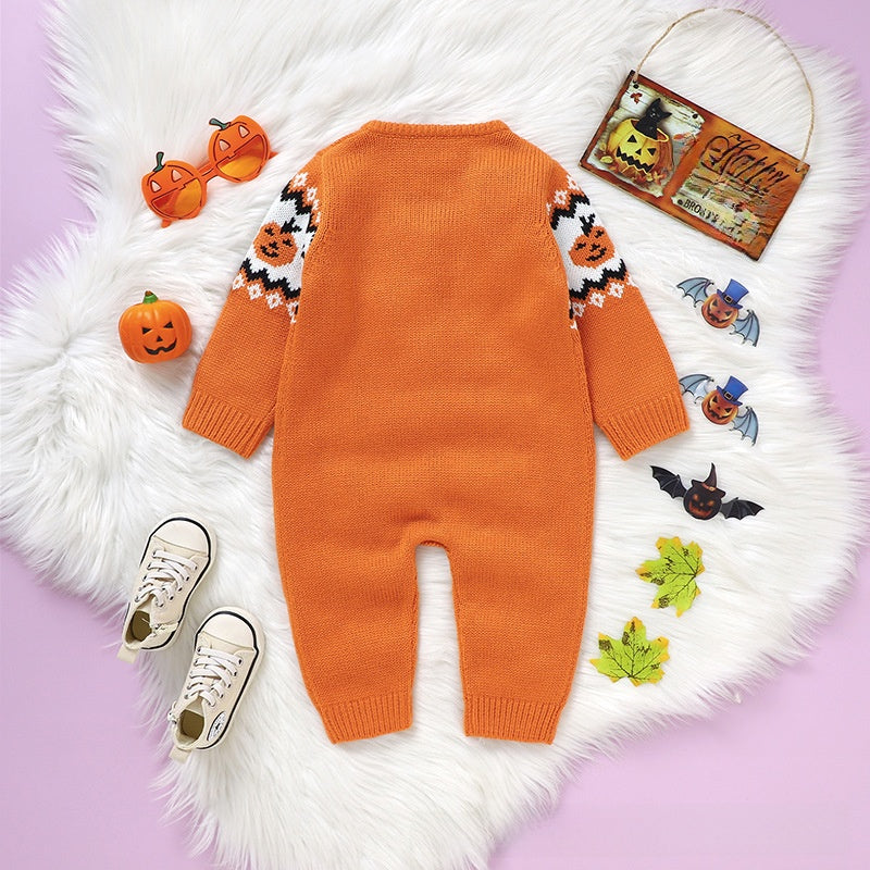 An orange knit baby jumpsuit with Halloween-themed patterns on the sleeves, laid flat on a white faux fur rug with small props including orange glasses, a pumpkin, shoes, and a hanging sign.