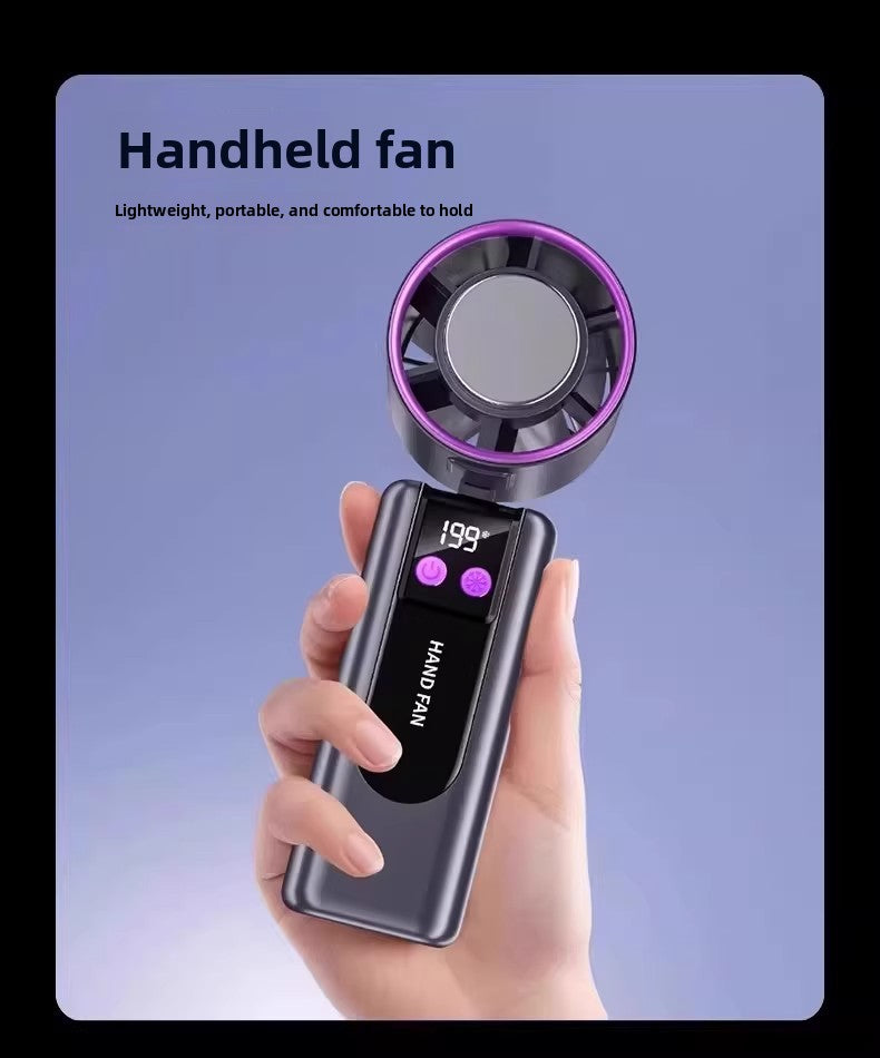 A hand holding a small handheld fan with a digital display showing 99 degrees and the words 'HAND FAN' on the front. The fan has a dark gray body with a glowing purple circular accent around the blades.