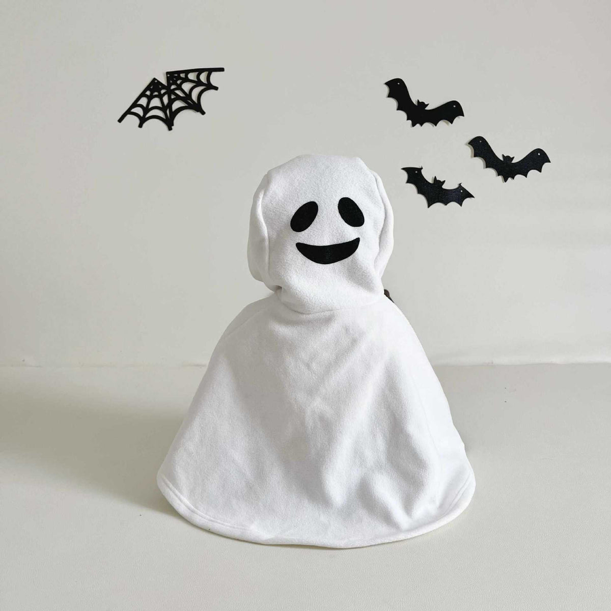 White ghost costume with black face design on a light gray background with Halloween decorations.