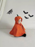 A baby wearing an orange pumpkin-shaped hooded cape with a small green stem, sitting on a white studio floor against a white wall with black bat and spiderweb decorations.