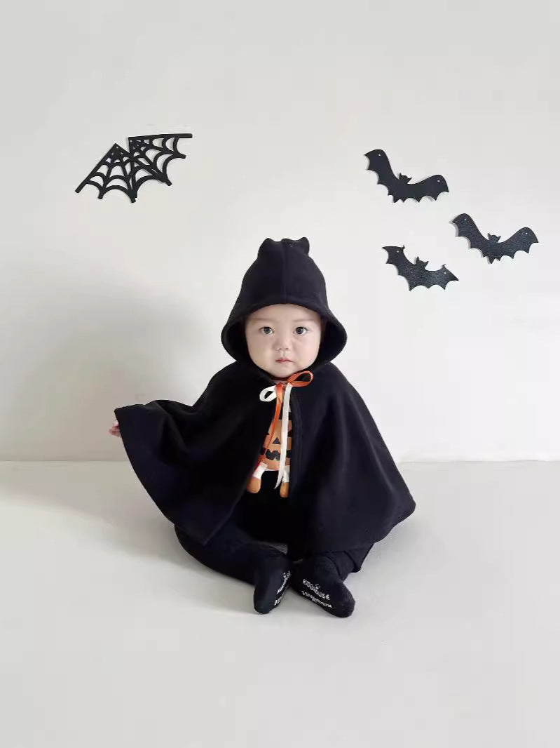A baby wearing a black hooded cape costume with a white and orange spider print neck scarf, sitting on a white floor against a white wall decorated with black bat and spiderweb cutouts.